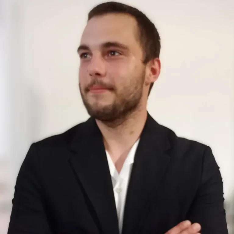 Homme en costume noir avec les bras croisés, regard sérieux et fond clair.