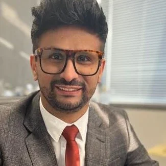 Homme en costume avec des lunettes, souriant à l'intérieur d'un bureau.